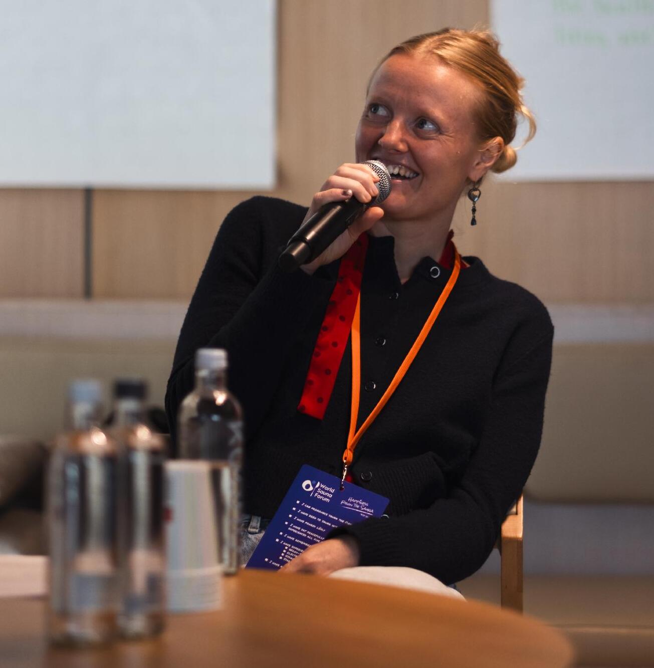 Polly Wilson, sat at a table with a hand held microphone, speaking at World Sauna Forum 2026