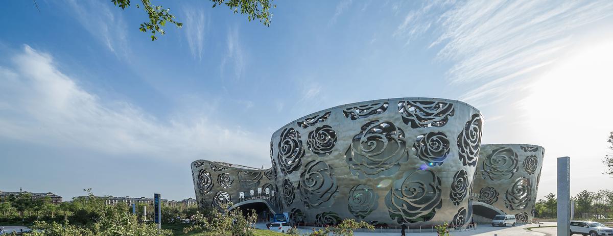 The museum is a solid volume covered by a detached silk-like skin of stainless steel with a pattern of cut-out Chinese roses / Xiao Kaixiong
